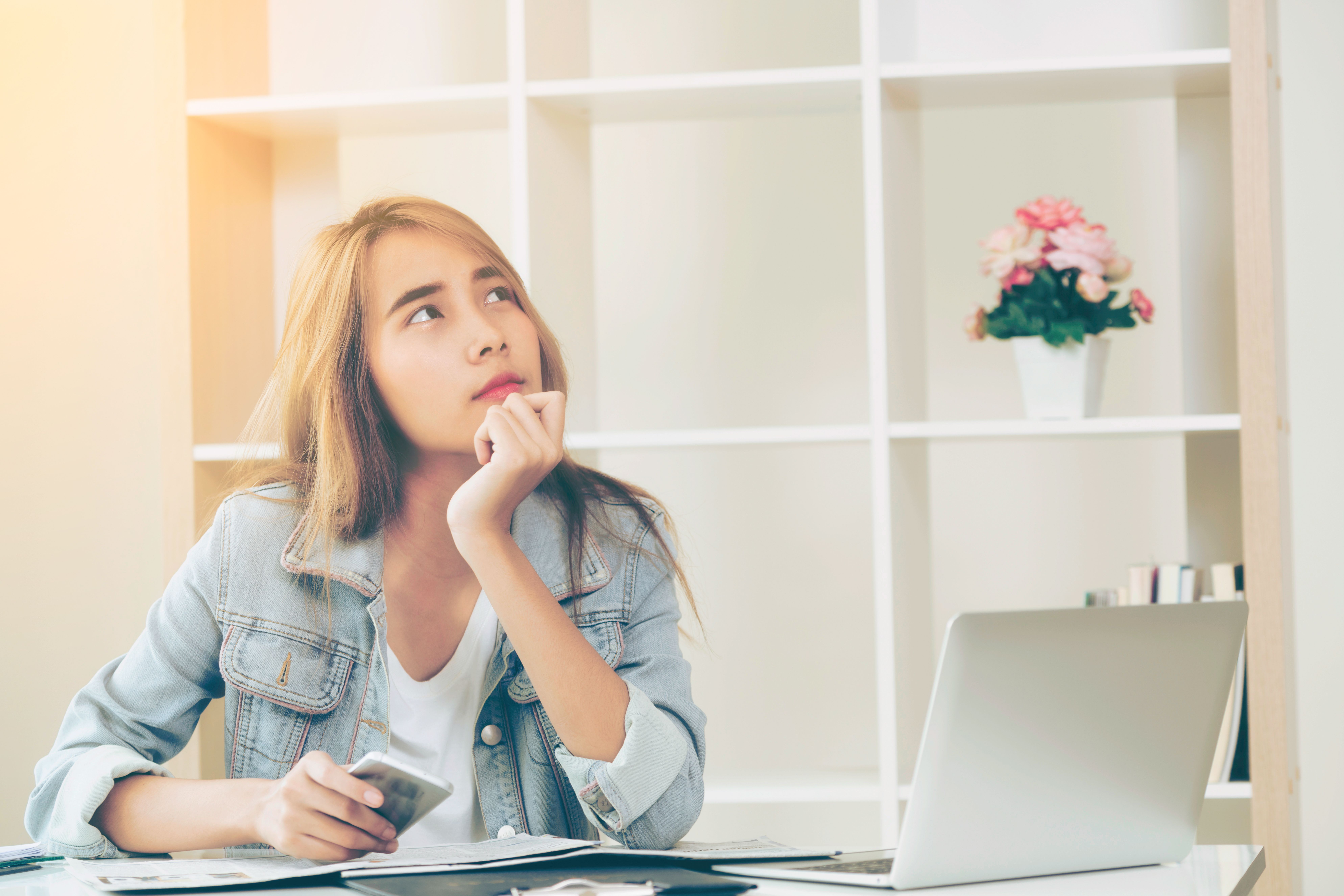Young woman in a denim jacket holds a smartphone and gazes upward thoughtfully at her desk beside a laptop.