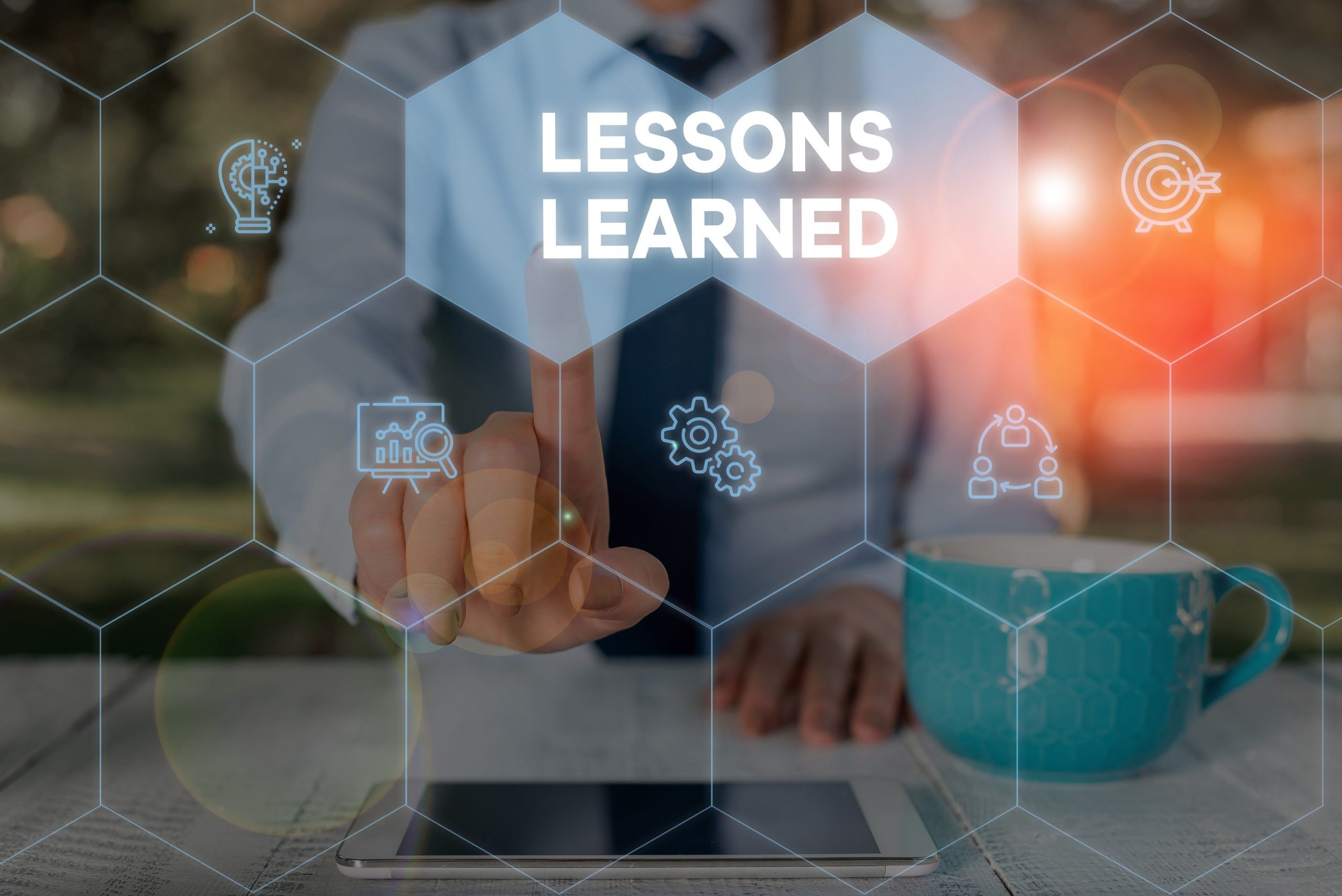 Businessperson touching a digital "Lessons Learned" interface with hexagonal icons, beside a teal coffee mug and tablet.