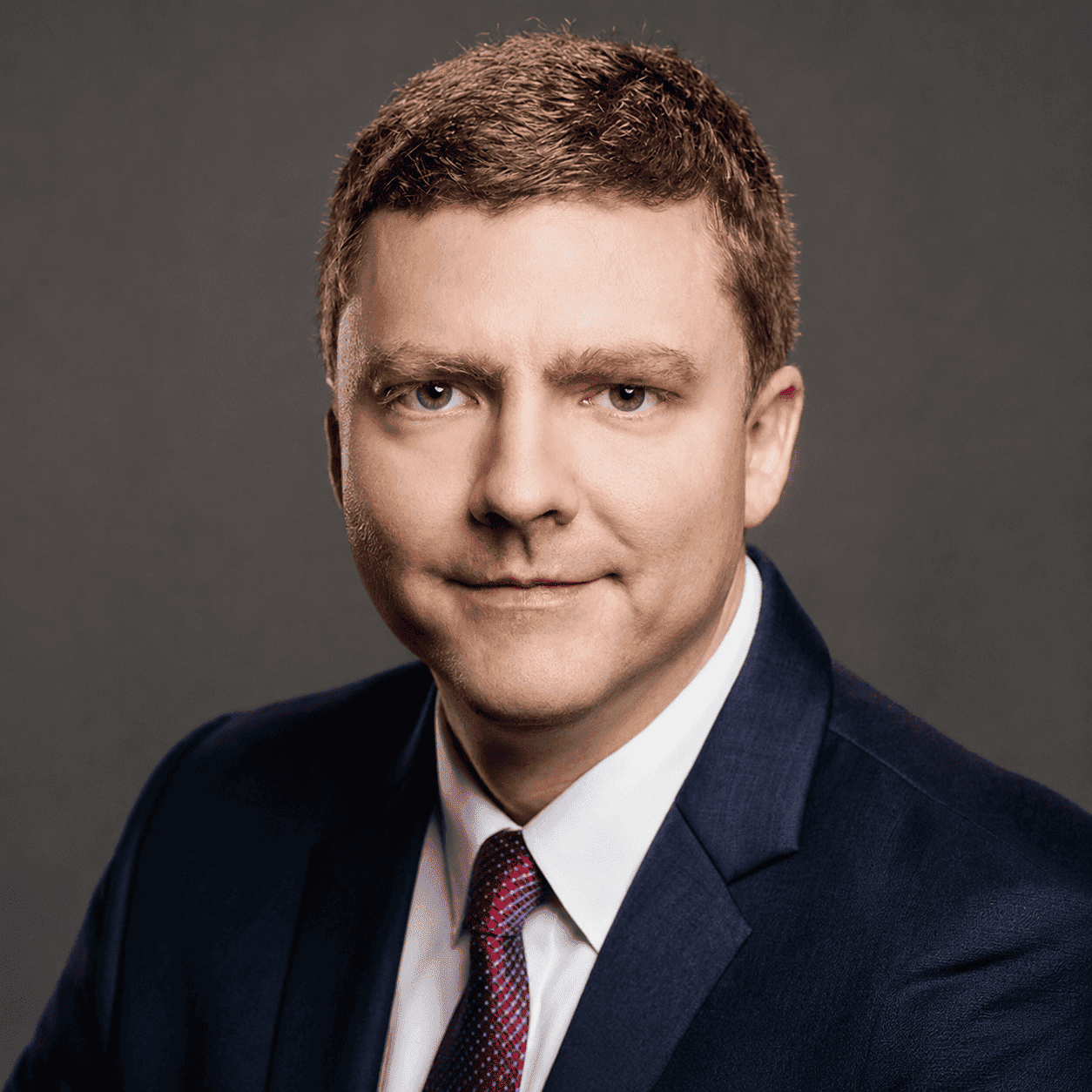 Professional headshot of Matt Hartley wearing a navy suit, white shirt, and patterned red tie against a gray backdrop.