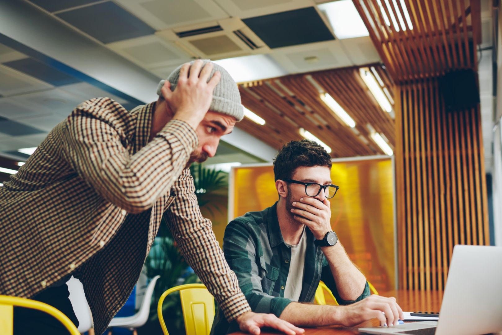 Two men with worried expressions stare at a laptop screen in a modern office, one clutching his head in distress.