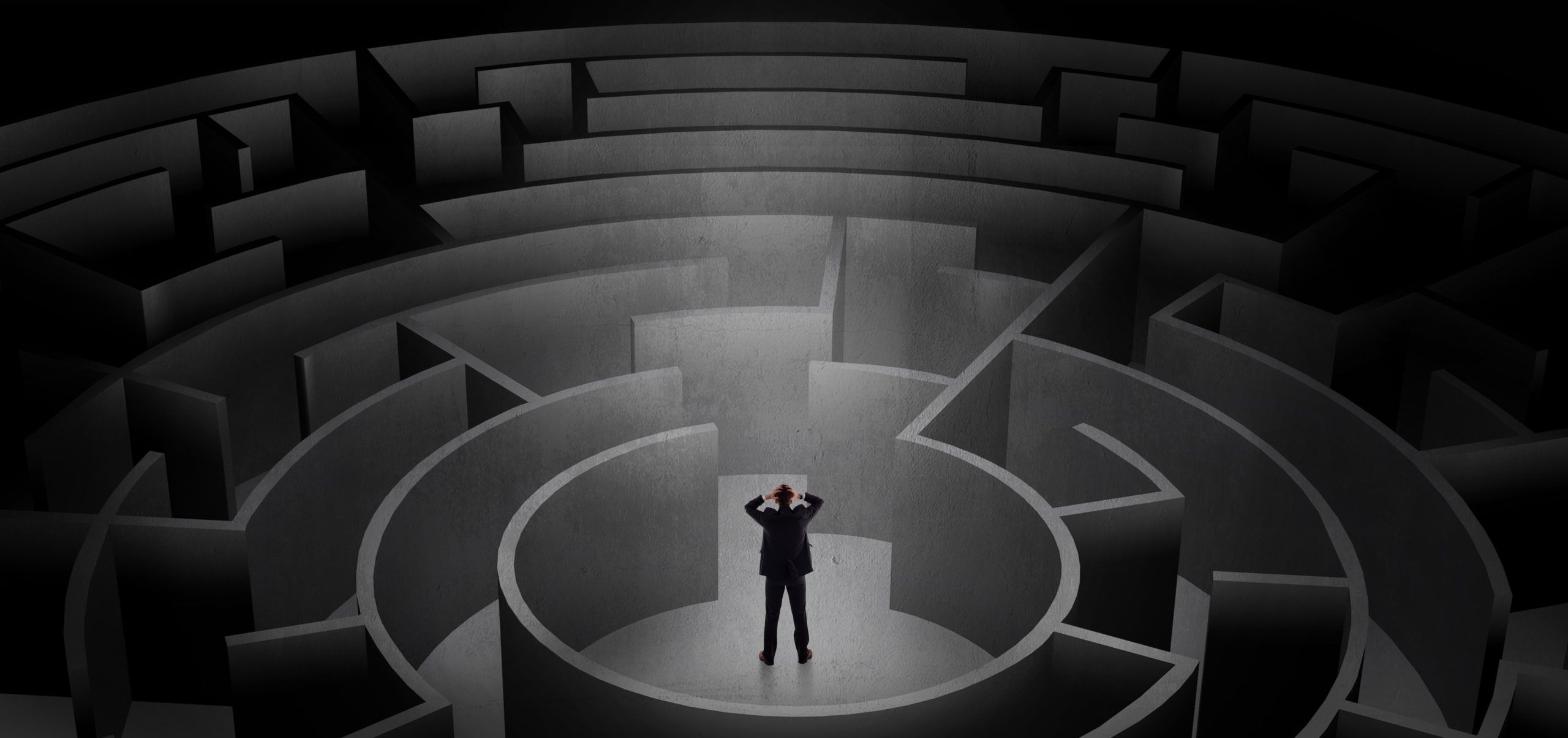 Businessman in a suit stands at the center of a vast dark circular maze, holding his head while facing multiple path choices.