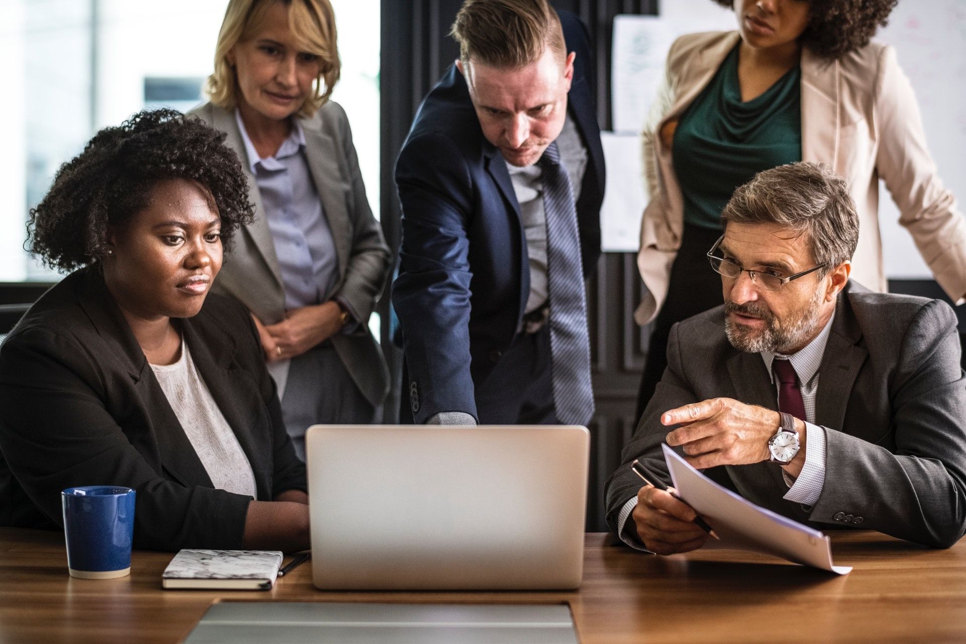 Diverse group of business professionals collaborating around a laptop during an office meeting with documents and coffee.