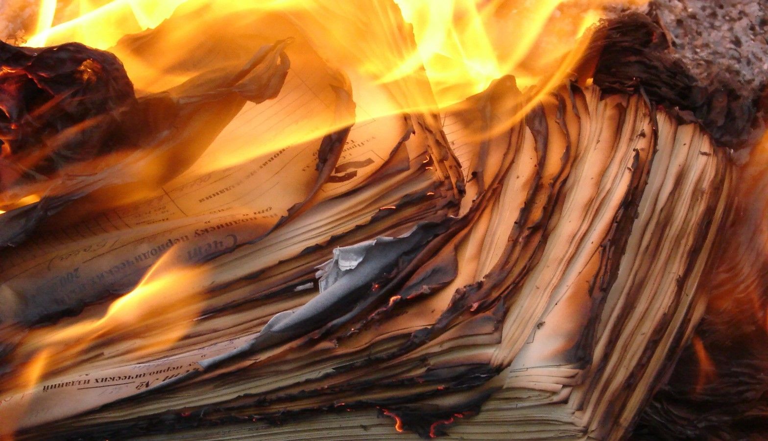 Stack of papers or documents engulfed in flames, with charred edges and curling pages amid bright orange fire.