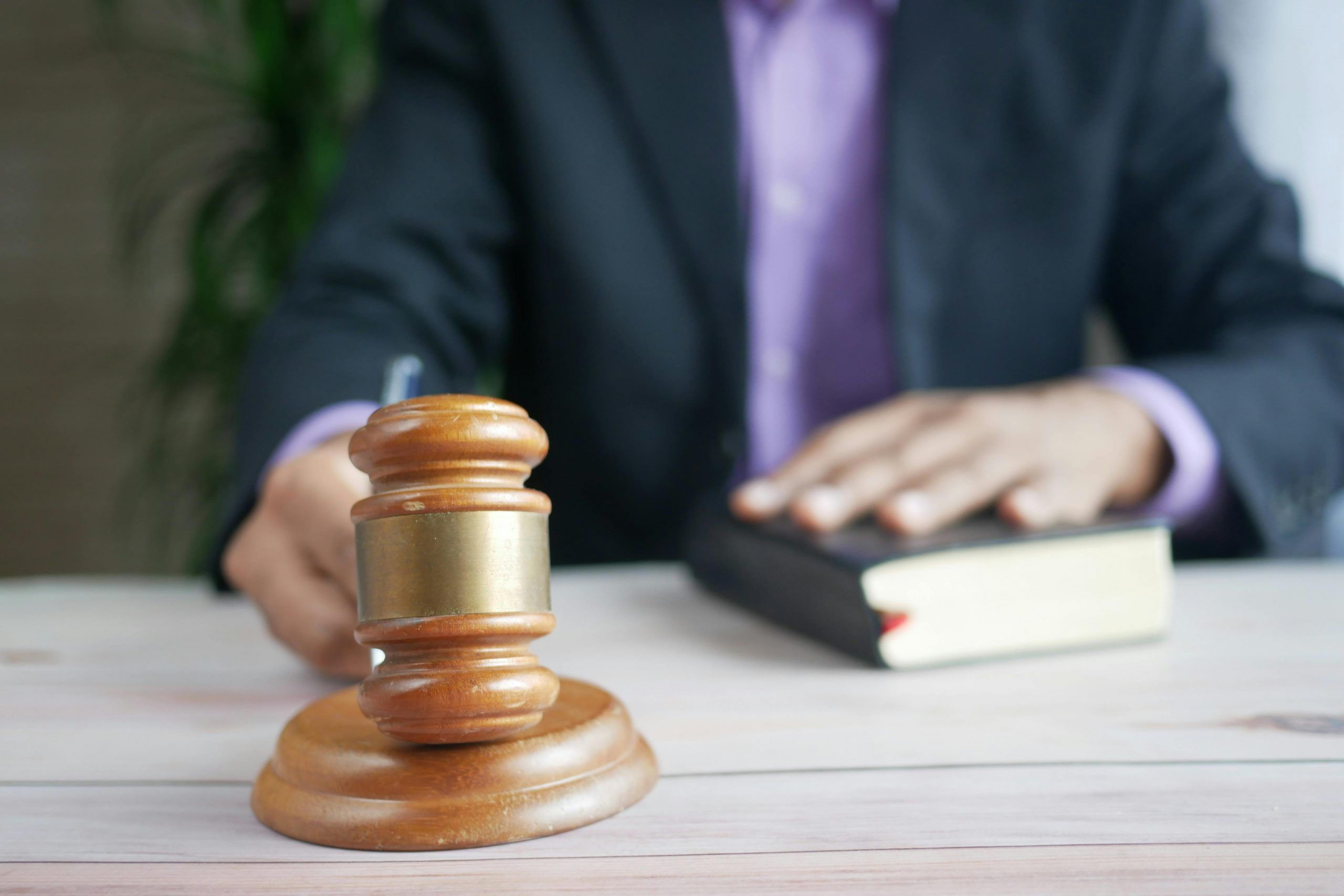 Wooden gavel on a desk with a lawyer in a dark suit resting his hand on a law book in the background.