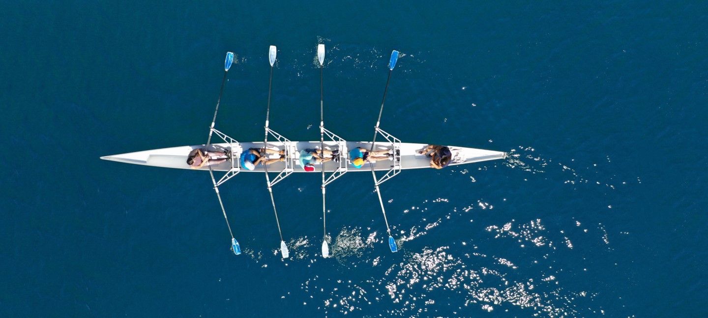 Aerial view of four rowers synchronizing their oars in a sleek racing scull on deep blue water.