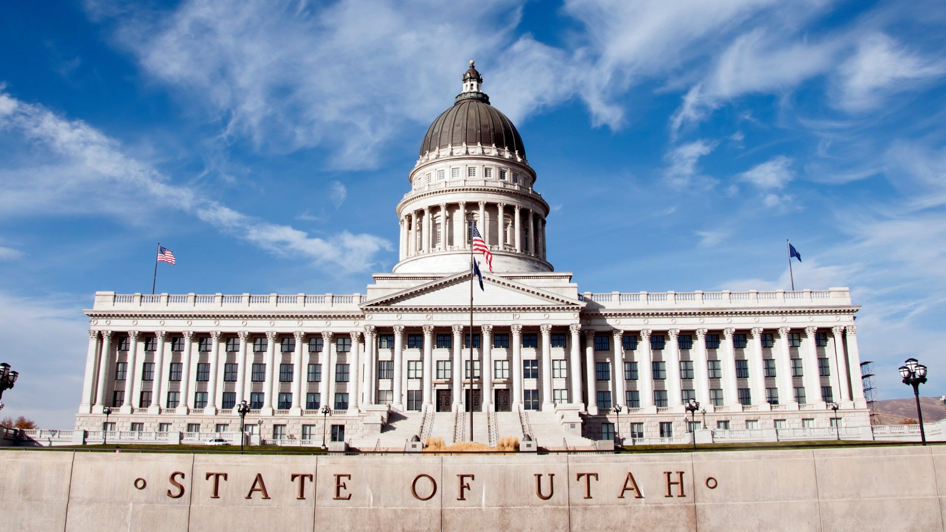 Utah State Capitol building with its grand domed roof, columned facade, and flags against a bright blue sky.